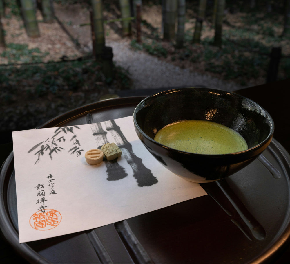 A black ceramic bowl filled with green matcha tea sits on a dark round tray next to a white paper card with black brushstroke artwork and a small green tea biscuit, with a blurred bamboo forest in the background.